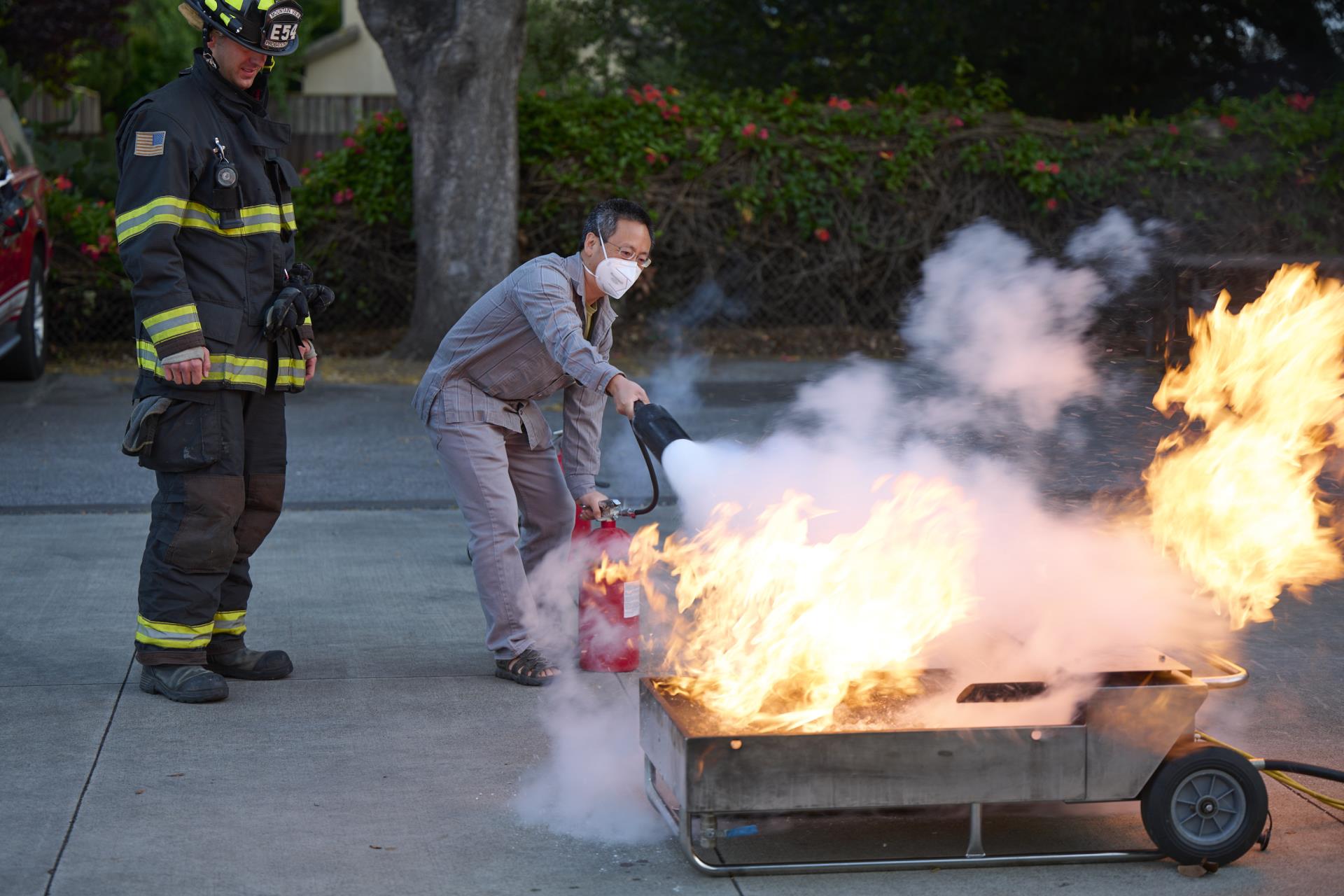 Person putting out fire with fire extinguisher while firefighter supervises